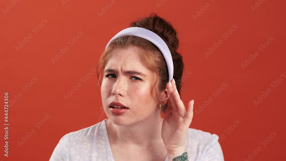 A redhead girl in a dress and with a rim on her head doing don�t hear gesture in an orange studio
