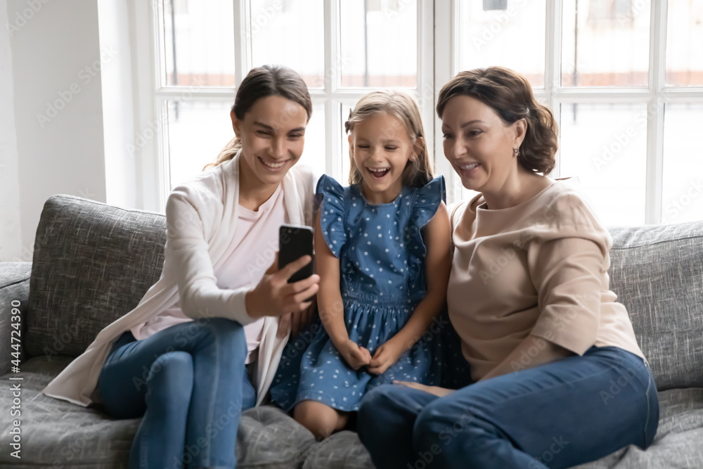 Overjoyed three generations of women relax on sofa at home laugh talk speak on video call on cellphone together. Happy little girl with mom and grandmother use smartphone have webcam virtual event.