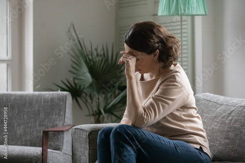 Upset mature Caucasian woman sit on couch feel depressed sad mourn miss or yearn in living room. Unhappy middle-aged female think or ponder of life or health problems. Maturity, loneliness concept.