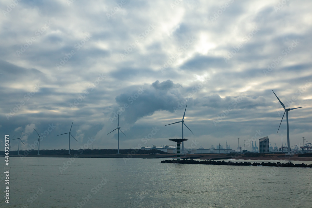 Wind farms onshore along a water channel in the Netherlands. Wind turbines by the water on the shore. Wind farms onshore along a water channel in the Netherlands. Wind turbines by the water on the shore.