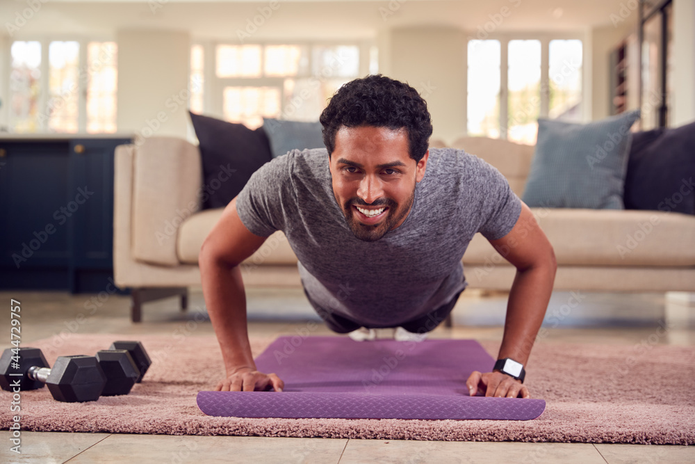 Fototapeta premium Man In Fitness Clothing At Home In Lounge Doing Press Ups And Exercising With Hand Weights