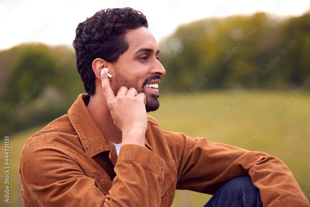 Man Sitting On Bench In Countryside Relaxing And Listening To Music Or Podcast On Wireless Earphones Man Sitting On Bench In Countryside Relaxing And Listening To Music Or Podcast On Wireless Earphones