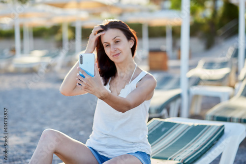 Young brunette with a phone on a sunbed on the beach. Vacation concept, taking a selfie, chatting with friends or freelancer, working remotely, webinar, communication, learning concept.