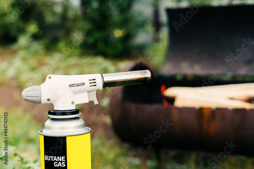 Close up man holds a gas burner with a balloon and makes a fire in the grill. Flame gun on the background of green grass in the evening in the back yard. Men hands light a fire.