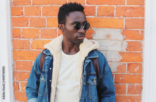 Portrait of stylish young african man model wearing denim jacket posing on a city street over brick wall background