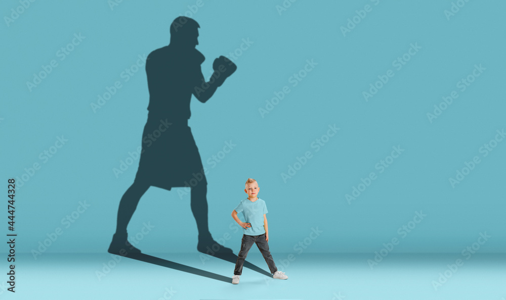 Childhood and dream about big and famous future. Conceptual image with boy and shadow of strong male boxer on blue background. Dreams, imagination, education concept. Childhood and dream about big and famous future. Conceptual image with boy and shadow of strong male boxer on blue background. Dreams, imagination, education concept.