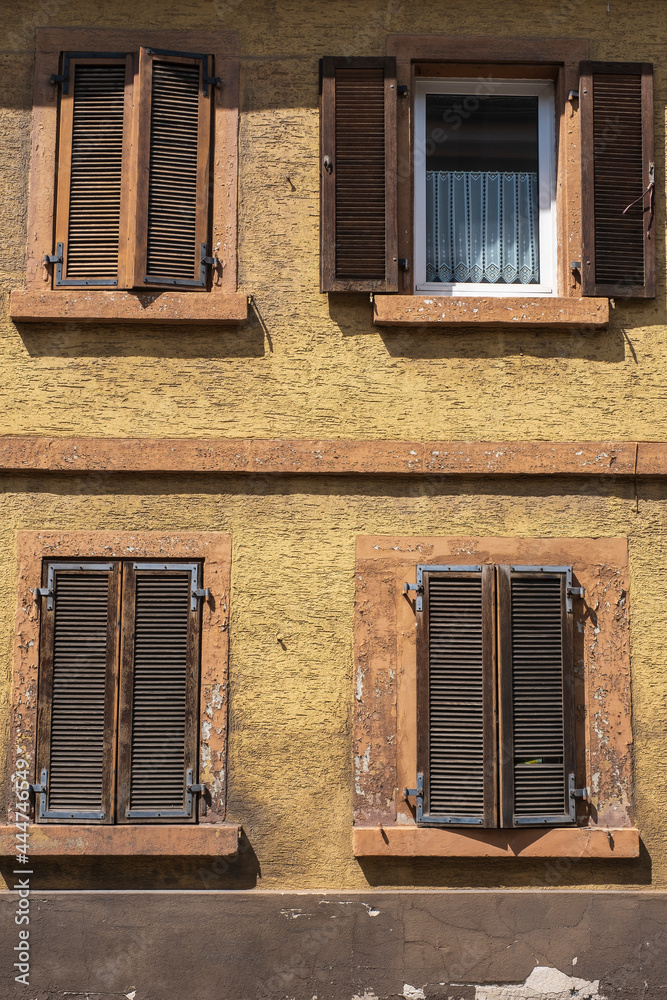 View of a weathered brown house facade with four windows, three with shutters 