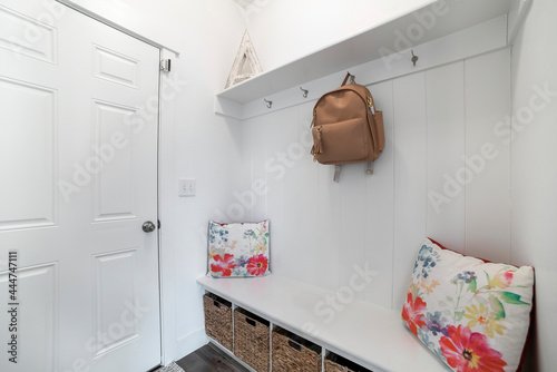 Interior of a white mud room with white door, built-in white bench and wall mounted hooks and shelf