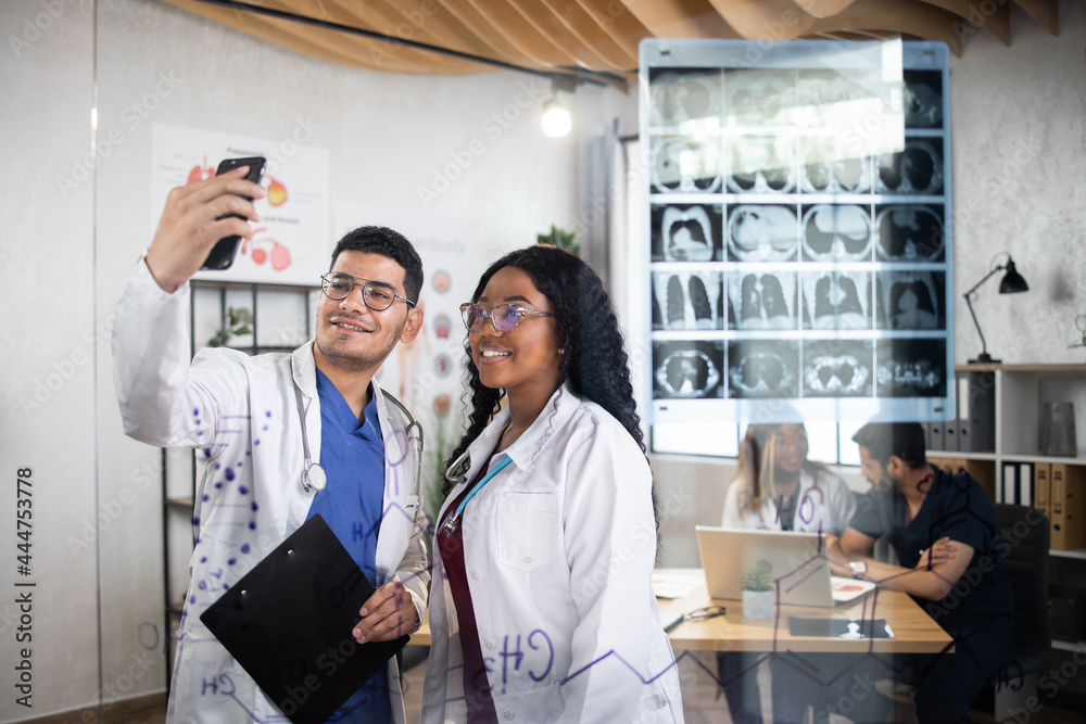 Fototapeta premium Smiling african woman and indian man in white lab coat taking selfie on smartphone. Two medical workers making photo after international meeting. View through the glass wall