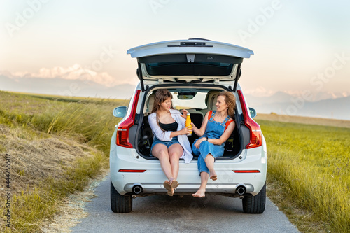 Mother and teenager daughter enjoying their road trip in countryside sitting in open car boot having drink. Local traveling, summer holidays lifestyle.