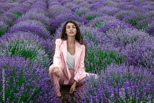 Lovely young woman wearing light pink business suit and sitting in purple lavender field; professional retouch.