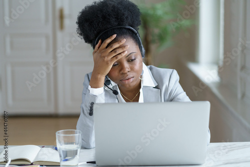 Foto Tired african american female support service or call center worker wearing wireless headset frustrated touching forehead speaking to client look upset annoyed on laptop screen