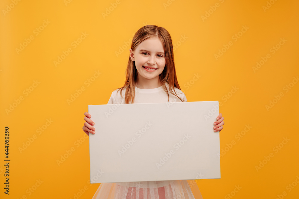 Pretty smiling girl child holding white canvas and looking at the camera isolated on yellow background with copyspace. Horizontal portrait of kid holding linen with place for text Pretty smiling girl child holding white canvas and looking at the camera isolated on yellow background with copyspace. Horizontal portrait of kid holding linen with place for text
