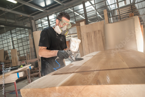 Man in respirator mask painting wooden planks at workshop. Craftsman modern furniture factory paint