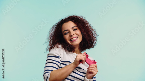 KYIV, UKRAINE - JUNE 30, 2021: happy young woman holding pink gamepad isolated on blue © LIGHTFIELD STUDIOS