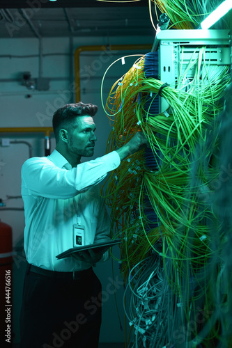 Vertical portrait of handsome data engineer inspecting cables in server room while working with supercomputer in blue light