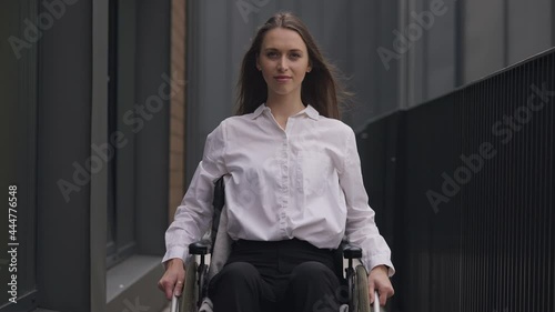Medium shot portrait of young gorgeous disabled Caucasian woman sitting in wheelchair looking at camera. Successful beautiful paraplegic businesswoman posing outdoors. Confidence and disability photo