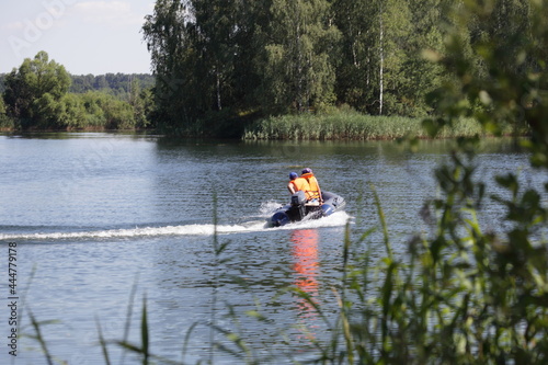 Moving away inflatable motor boat with outboard motor and man in orange lifejacket fast floating on the water on green trees background at Sunny summer day, beautiful European river landscape