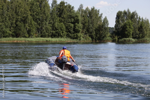 Moving away inflatable motor boat with outboard motor and man in orange lifejacket fast floating on the water on green forest background at Sunny summer day, beautiful Russian river landscape