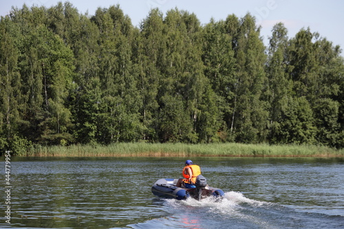 Go out inflatable motor boat with outboard motor and man in orange lifejacket fast floating on the water on green forest background at Sunny summer day, beautiful European lake landscape