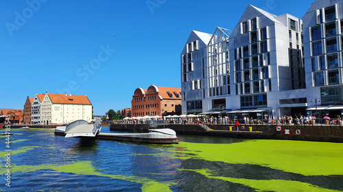 Gdansk, Poland - July 11, 2021: View of the old city of Gdansk on the Motlawa River. Tourists walk along the waterfront. On Motlawa green patches of duckweed. Middle of the river flows ship.