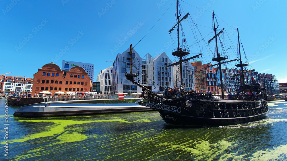 Obraz premium Gdansk, Poland - July 11, 2021: View of the old city of Gdansk on the Motlawa River. Tourists walk along the waterfront. On Motlawa green patches of duckweed. Middle of the river flows ship.
