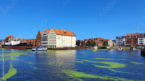 Gdansk, Poland - July 11, 2021: View of the old city of Gdansk on the Motlawa River. Tourists walk along the waterfront. On Motlawa green patches of duckweed. Middle of the river flows ship. © abrada