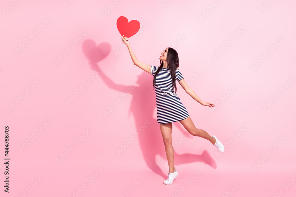 Full length body size view of pretty cheerful woman holding heart card having fun jumping isolated over pink pastel color background Full length body size view of pretty cheerful woman holding heart card having fun jumping isolated over pink pastel color background