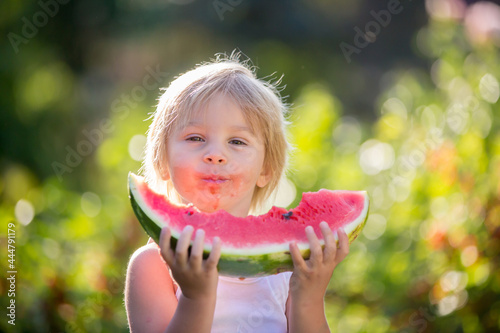 Cute blond child, toddler boy and pet dog, eating watermelon in garden