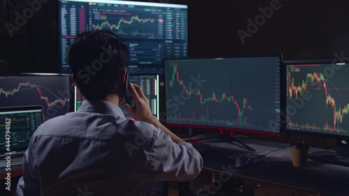 Back View Of A Asian Young Male Stock Market Broker Talking On Mobile Phone With Analyzing Graphs On Multiple Computer Screens
