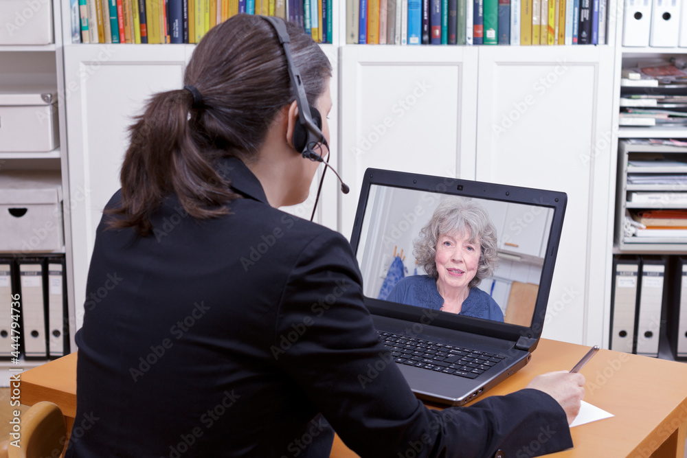 Female lawyer with headset in front of her laptop writing something on a paper while having a live video chat with an elderly client, copy space Female lawyer with headset in front of her laptop writing something on a paper while having a live video chat with an elderly client, copy space