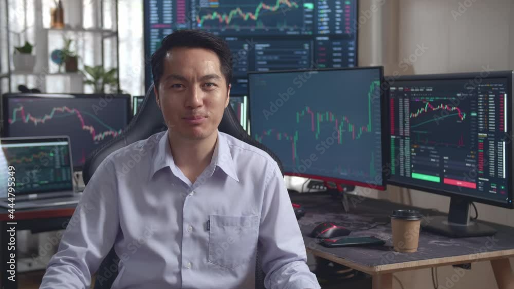 Asian Male Stock Market Broker Speech To Camera With Analyzing Graphs On Multiple Computer Screens At The Background
