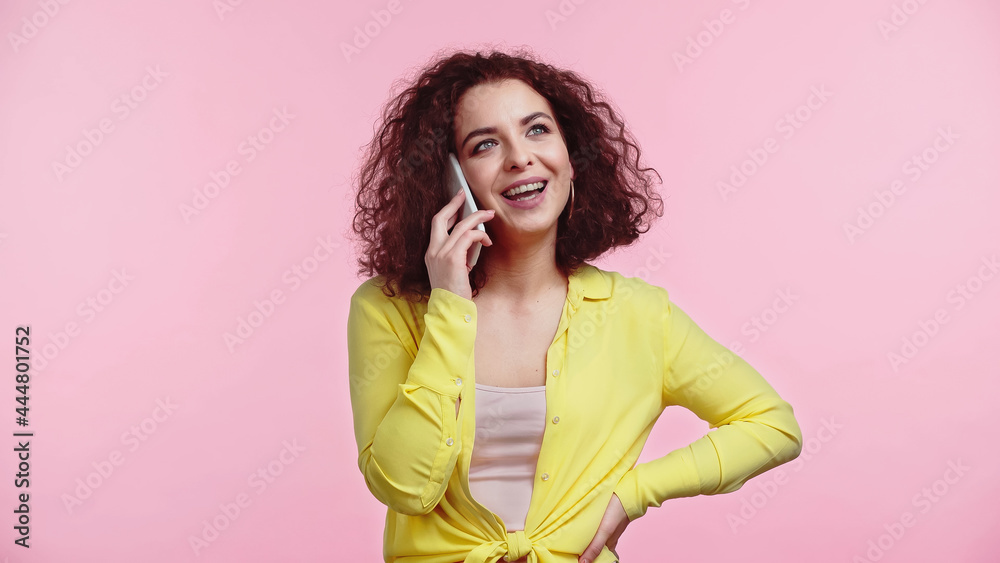happy woman standing with hand on hip while talking on cellphone isolated on pink happy woman standing with hand on hip while talking on cellphone isolated on pink