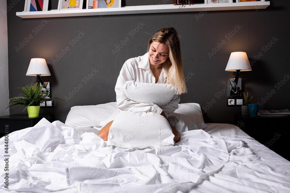 A beautiful girl is sitting in bed and hugging a pillow. In a white shirt, smiling A beautiful girl is sitting in bed and hugging a pillow. In a white shirt, smiling