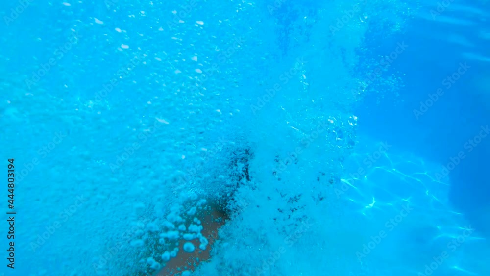 SLOW MOTION, UNDERWATER: Young boy speeding and jumping into swimming pool, kid doing a cannonball into clear water of a summer house. Courtyard of a summer house with wooden floor.