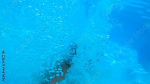 SLOW MOTION, UNDERWATER: Young boy speeding and jumping into swimming pool, kid doing a cannonball into clear water of a summer house. Courtyard of a summer house with wooden floor.