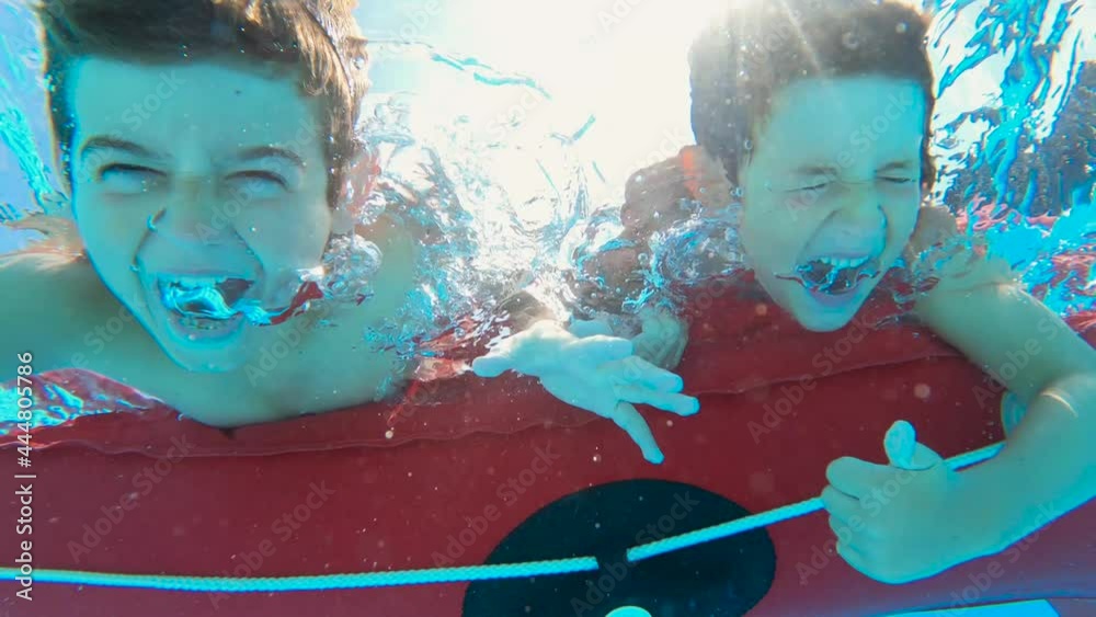 Two boys enjoying on a floating red boat in a pool, taking a deep breath to go underwater, looking at camera, smiling and screaming. Funny moments during summer vacation in family. Slow motion