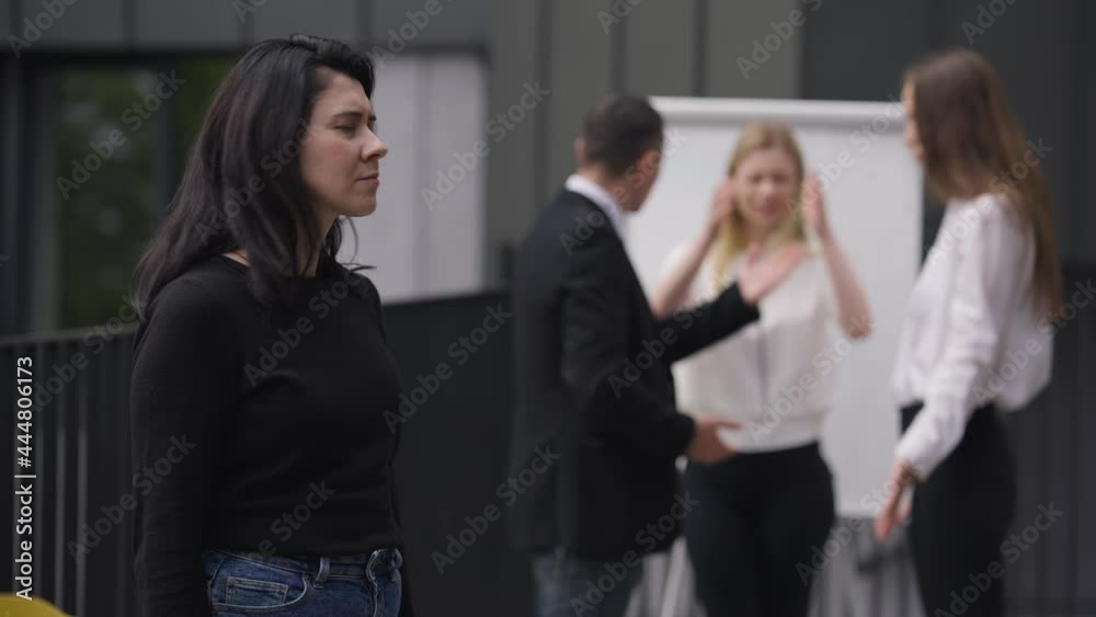 Caucasian woman closing ears with hands looking at camera as nervous colleagues arguing at background. Portrait of stressed employee posing outdoors with coworkers quarrelling. Inconvenience concept Caucasian woman closing ears with hands looking at camera as nervous colleagues arguing at background. Portrait of stressed employee posing outdoors with coworkers quarrelling. Inconvenience concept