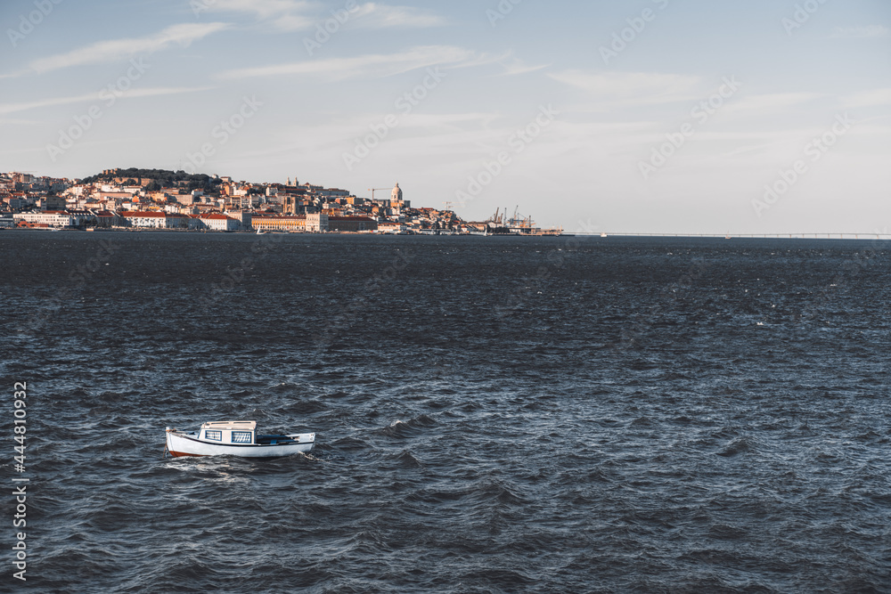 A small cozy boat with blue with striped curtains inside of cabin windows sways in the waves of Tagus river, Portugal, with a riverbank full of antique Lisbon houses of Alfama district in a background A small cozy boat with blue with striped curtains inside of cabin windows sways in the waves of Tagus river, Portugal, with a riverbank full of antique Lisbon houses of Alfama district in a background