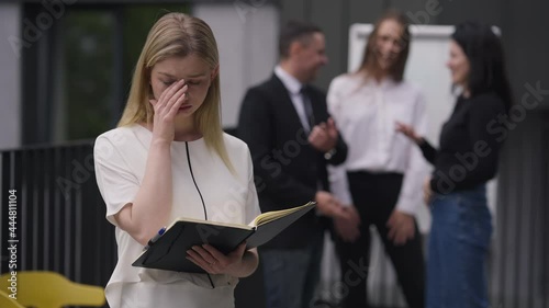 Stressed sad beautiful young woman crying standing outdoors with colleagues mocking at background. Caucasian men and women making fun of coworker at meeting outdoors. Stress and intolerance