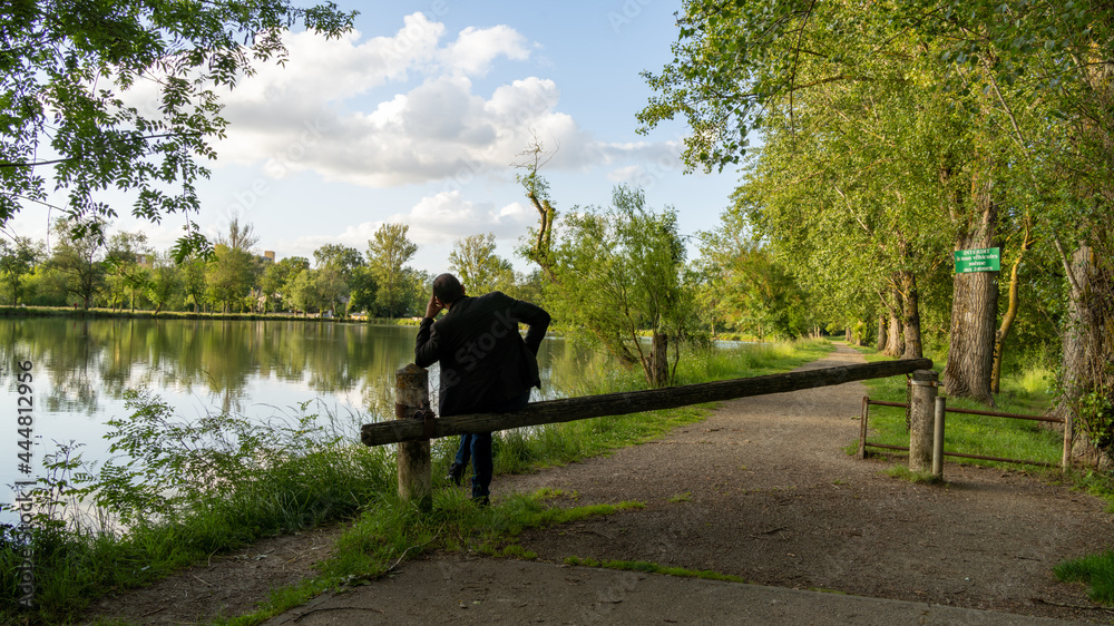 Stylish young man, from behind, sitting on a wooden fence, by a lake, on a late spring day Stylish young man, from behind, sitting on a wooden fence, by a lake, on a late spring day
