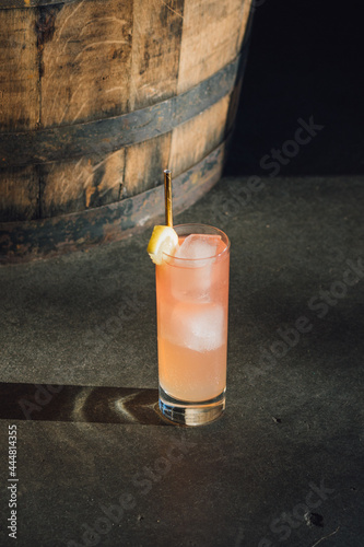 pink lemonade in tall Collins glass with lemon wedge and gold straw on concrete with whiskey barrel 