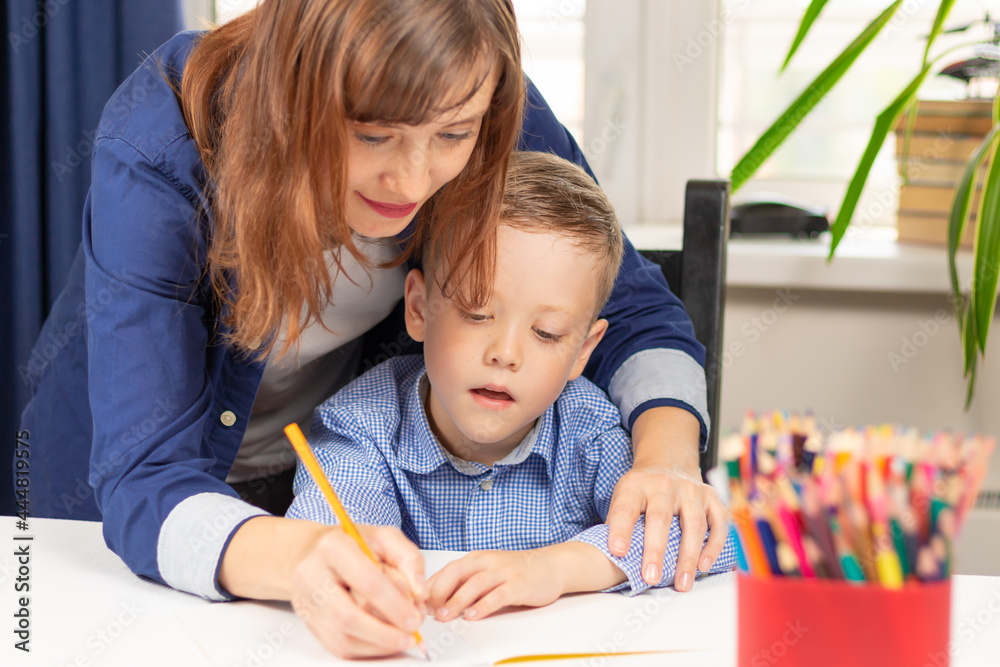 Cute seven year old baby boy doing homework at home with mom on a white wooden table during a pandemic. Selective focus. Close-up. Portrait Cute seven year old baby boy doing homework at home with mom on a white wooden table during a pandemic. Selective focus. Close-up. Portrait