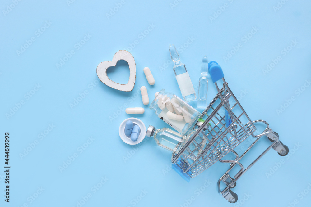 Shopping cart with medicines for online shopping with a wooden heart on a blue background. Tablets of vitamins and antibiotics with pain relievers, capsules, ampoules for influenza and colds. Shopping cart with medicines for online shopping with a wooden heart on a blue background. Tablets of vitamins and antibiotics with pain relievers, capsules, ampoules for influenza and colds.