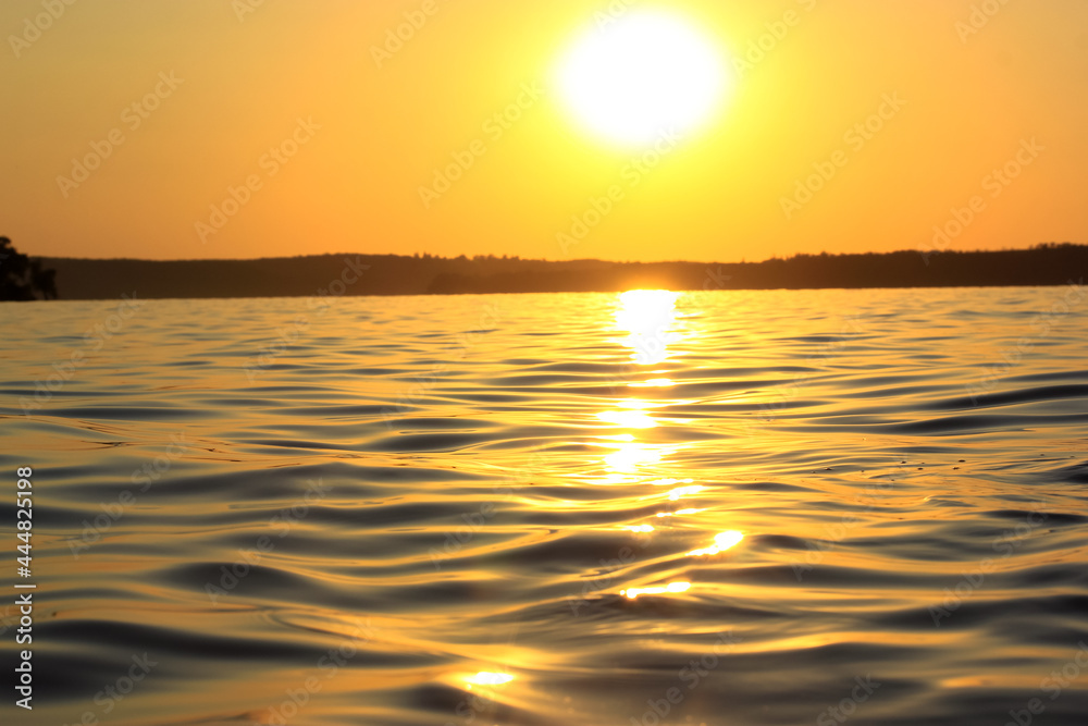 Sun at sunset and sea waves close up, low-angle of view. Water surface with small waves. River, lake, pond. Huge round sun in orange sky on a horizon. Golden rays of sun reflected on surface of water.