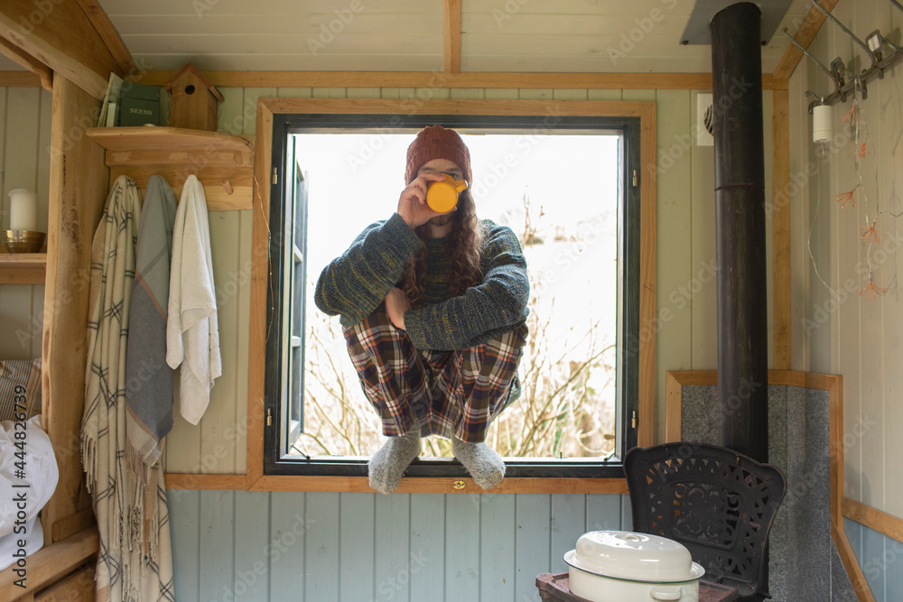 © Sam Edwards/Caia Image - Young man in pajamas drinking coffee in tiny cabin window