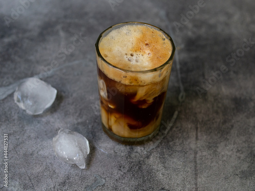 Ice coffee in a tall glass, ice cubes on a gray wooden table. Cold summer drink with tubes on a black background with copy space. Close up.
