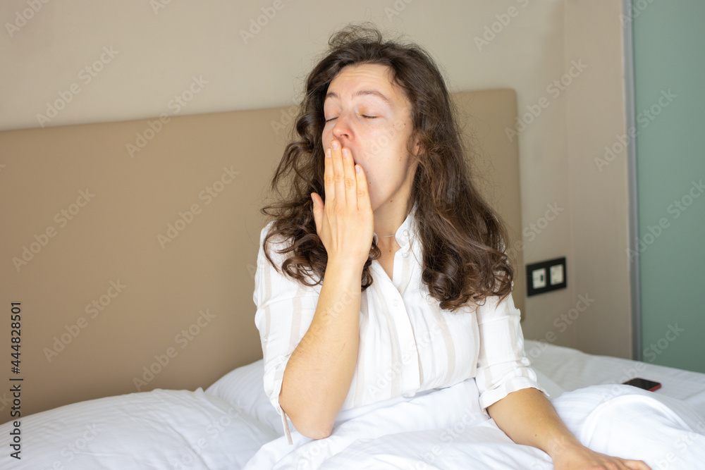Sleepy exhausted young brunette woman waking up in white bed at home, yawning covering her mouth, feeling headache or migraine, hangover. Suffering from insomnia, lack of sleep in morning. Copy space Sleepy exhausted young brunette woman waking up in white bed at home, yawning covering her mouth, feeling headache or migraine, hangover. Suffering from insomnia, lack of sleep in morning. Copy space