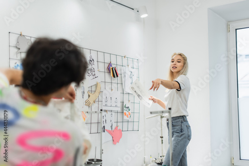 blonde fashion designer pointing with finger near sketches and blurred colleague