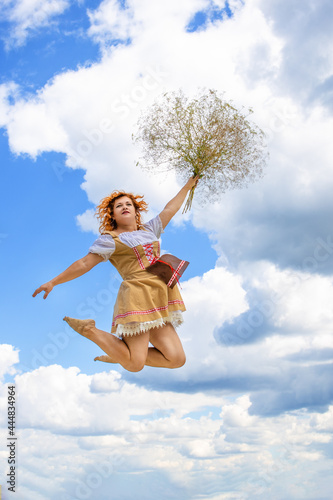Girl with red hair jumping against the background of clouds with a bouquet of field herbs